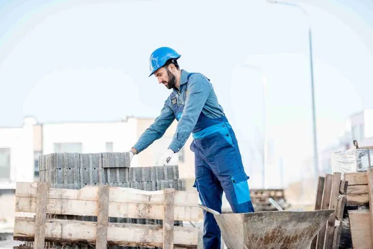 Construction worker wearing a blue hard hat and gloves stacking concrete paving stones on a pallet at an outdoor job site, with tools and materials nearby.